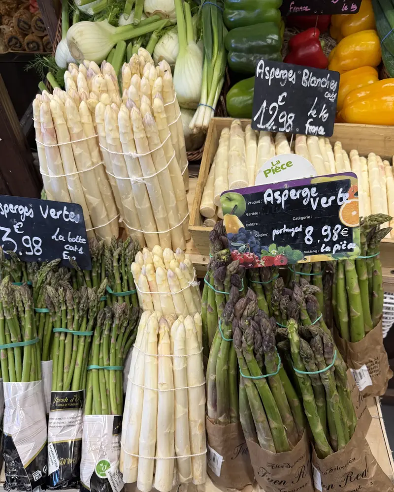 Bottes d'asperges blanches et vertes en cagette, avec leurs prix sur des ardoises, devant les poivrons et les fenouils