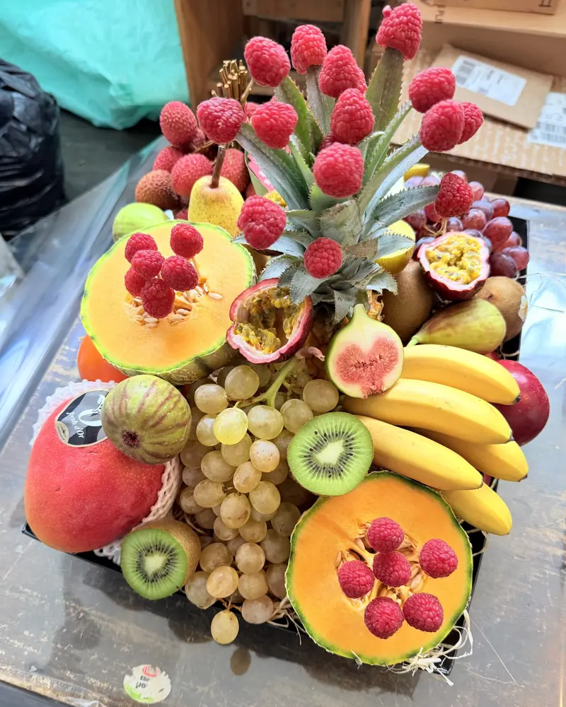 Corbeille de fruits : ananas hérissé de framboises, demi-melons, figues, fruits de la passion, raisin, bananes, kiwis et mangue