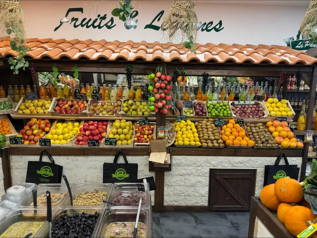 L'intérieur de la boutique : étals sous un petit toit de tuiles, cagettes de pommes, poires, agrumes et kiwis, bouteilles de jus, bar à olives